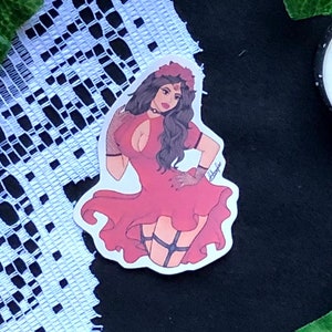 Könnte beinhalten: Ein Aufkleber mit einer Frau mit roten Haaren, die ein rotes Kleid trägt und eine rote Rose hält. Der Aufkleber befindet sich auf einem schwarzen Hintergrund mit weißer Spitze und Kerzen.