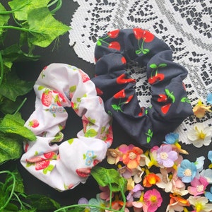 Könnte beinhalten: Zwei Stoff-Haargummis, eines schwarz mit weißen Kirschen und eines weiß mit roten Erdbeeren. Beide Haargummis liegen auf einem weißen Spitzenhintergrund mit rosa und weißen Blumen.