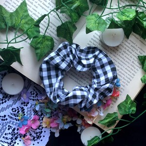 May include: Black and white checkered scrunchie hair tie. The scrunchie is made of fabric and has a gathered look. It is lying on a surface with flowers and candles.