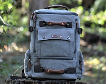 May include: A gray canvas backpack with brown leather accents and a zippered front pocket. The backpack has a top handle and adjustable shoulder straps.