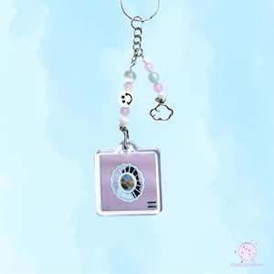May include: A clear acrylic keychain with a square pendant featuring a light purple album cover design. The keychain includes a silver chain, smiley face beads, star charm, cloud charm, and pink and blue beads.
