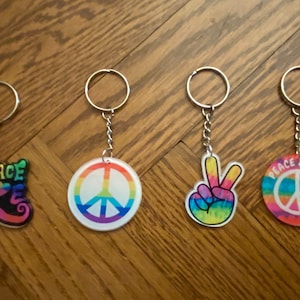 May include: Four keychains with peace symbols and phrases. One keychain has the word "PEACE" in a colorful, psychedelic font. Other keychains feature a peace sign, a peace hand sign, and the words "PEACE & LOVE".