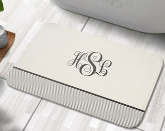 Puede incluir: Una alfombra de baño blanca con un borde rayado en blanco y negro y un monograma de las letras "HSL" en una fuente de letra cursiva gris.