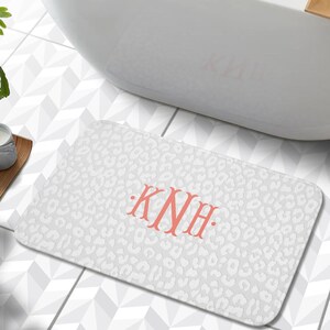 May include: White bath mat with a leopard print design and the monogram "KNH" in coral pink.