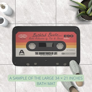K&ouml;nnte beinhalten: Eine schwarze und graue Badematte in Form eines Kassettenbandes. Das Band tr&auml;gt den Text "Bathtub Beats (2001)" und "Retro Relaxation by Tina & Steven". Die Matte ist 86 cm x 53 cm gro&szlig;.