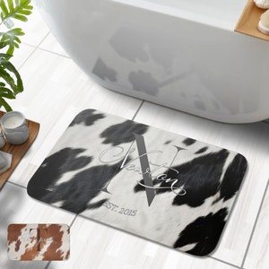 Puede incluir: Una alfombra de baño de piel de vaca blanca y negra con una letra "N" gris y el texto "Newton EST. 2015" en blanco.