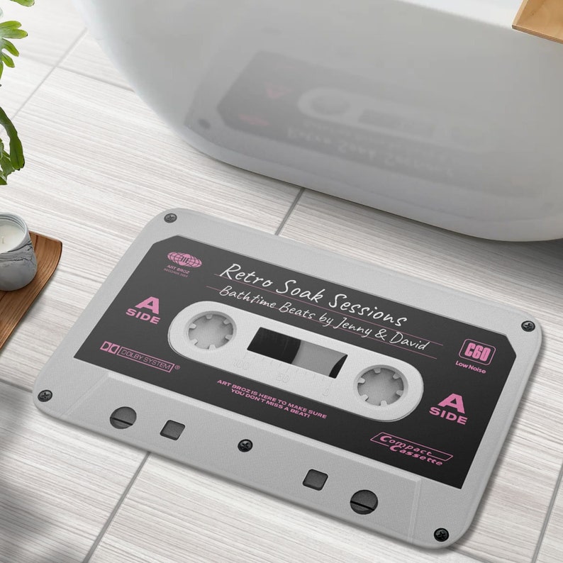 K&ouml;nnte beinhalten: Eine graue und schwarze Badematte in Form einer Kassette. Die Matte tr&auml;gt den Text "Retro Soak Sessions" und "Bathtime Beats by Jenny & David". Die Matte tr&auml;gt auch den Text "A Side" und "C60 Low Noise".