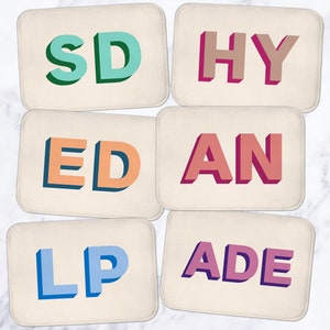May include: Six rectangular beige coasters with colorful letters spelling out the word "SHY LADE AN ED".
