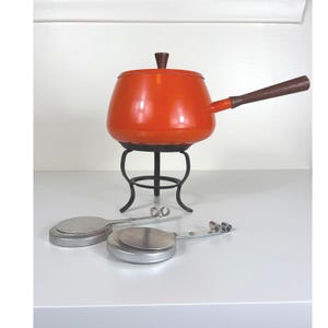May include: A bright orange fondue pot with a dark brown wooden handle and matching lid knob, resting on a black metal stand. Two circular silver metal fuel burners with extended handles are positioned in front of the stand on a flat surface.