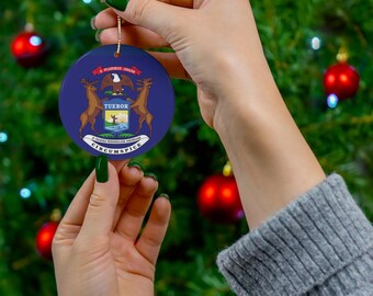 Puede incluir: Un adorno navideño redondo azul y blanco con el sello del estado de Michigan. El sello representa un águila calva con las alas extendidas, un escudo con una salida del sol y un hombre sosteniendo un arco y una flecha, y las palabras "Tuebor" y "E Pluribus Unum".