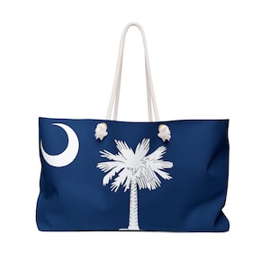 Puede incluir: Una bolsa de lona azul y blanca con el diseño de la bandera del estado de Carolina del Sur. La bolsa tiene asas de cuerda blanca y botones de madera.