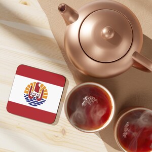 May include: A copper-coloured teapot and two cups of tea are on a light-coloured wooden surface. A square coaster with the flag of French Polynesia is also present. The tea is steaming, suggesting it is freshly brewed.