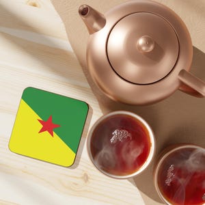 Op de afbeelding: Een koperkleurige theepot en twee kopjes thee staan op een licht houten oppervlak. Er is ook een vierkante onderzetter met de vlag van Frans-Guyana. De theekopjes zijn gevuld met een donkere vloeistof en er komt stoom uit.