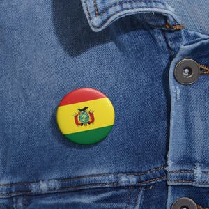 May include: A round button pin featuring the Bolivian flag, with red, yellow, and green colors. The pin is affixed to a blue denim jacket. The flag design includes a coat of arms in the center.