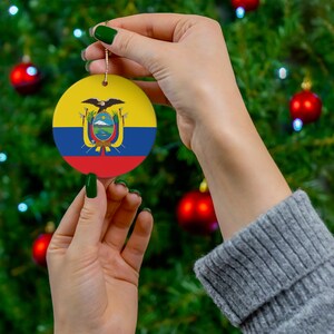 May include: A round Christmas bauble featuring the flag of Ecuador. The bauble is yellow, blue and red with the Ecuadorian coat of arms in the centre. The bauble is being held by a hand with green nail polish.