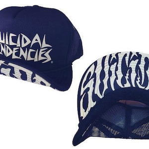 以下が含まれることがあります： 白のスタイリッシュな文字で「SUICIDAL TENDENCIES」と書かれたネイビーブルーのトラッカーハット。つばは白と黒の模様です。帽子はメッシュバックです。