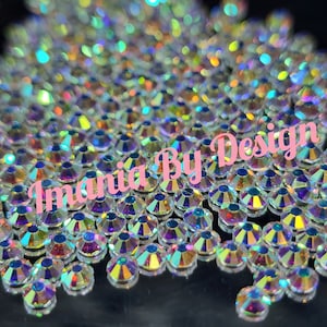 May include: A close-up of a pile of iridescent clear rhinestones. The rhinestones are small and have a faceted design. The rhinestones are scattered on a black surface. The text "Imania By Design" is visible in the image.