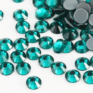 May include: A scattering of teal green rhinestones on a white background. The rhinestones are round and have a faceted surface.