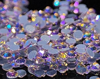 May include: A close-up of a pile of iridescent purple rhinestones. The rhinestones are scattered on a black surface and have a variety of sizes and shapes.