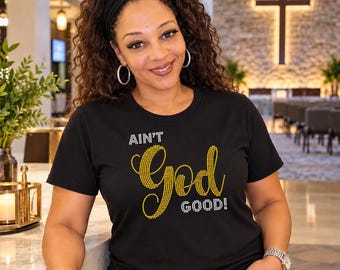 May include: A black short-sleeved t-shirt featuring the text AIN'T God GOOD! in yellow and white rhinestones. The word God is written in a large, cursive yellow font, while the words AIN'T and GOOD! are in a smaller white block font.