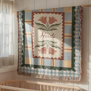 May include: A woven blanket with a floral design and the name "Heidi" in script. The blanket features a central rectangular design with red flowers and green leaves, framed by a scalloped border and a blue and white gingham pattern. The blanket is hanging on a wooden rod.