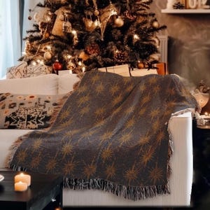 May include: A dark gray throw blanket with a repeating gold starburst pattern is draped over a white sofa. The blanket has a fringed edge. A Christmas tree with gold ornaments and lights is in the background.
