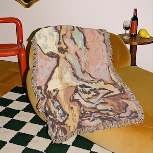 May include: A woven throw blanket featuring an abstract marbled pattern in shades of brown, tan, muted green, and soft pink. The rectangular blanket has a fringed edge and is draped over a curved yellow chair. Text at the bottom reads: THIS IS A MOCK UP, FINAL PRODUCT RESULTS MAY VARY.