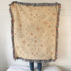 Peut inclure: Une couverture tissée beige avec un motif céleste d'étoiles et de nuages dans des tons de marron et de bleu. La couverture a des bords frangés multicolores. La couverture est présentée contre un mur blanc.