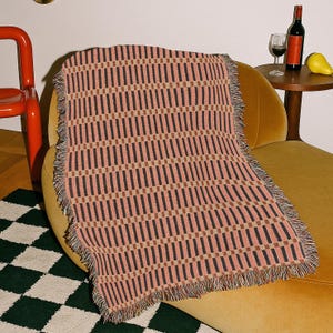 Könnte beinhalten: Eine gewebte Decke mit einem geometrischen Muster aus vertikalen schwarzen und braunen Streifen auf rosa Hintergrund. Die Decke hat einen Fransenrand und ist über einem gelben Sessel drapiert. Ein roter Stuhl und ein karierter Teppich sind im Hintergrund zu sehen.