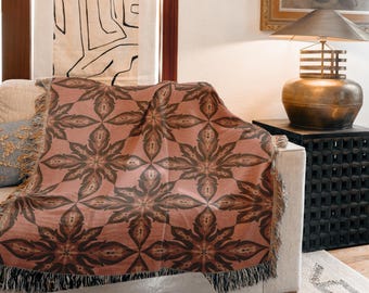 Op de afbeelding: Een geweven sprei met een herhalend bloemmotief in tinten van stoffig roze en bruin, gedrapeerd over een beige bank. De sprei heeft een gefranjerde rand. De kamer heeft ook een lamp en kunstwerken.