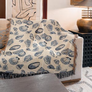 May include: A beige throw blanket with a repeating pattern of blue seashells. The blanket is draped over a sofa and has a fringed edge. The seashells vary in size and shape, creating a coastal theme.