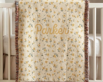 May include: A cream-colored baby blanket with a floral pattern of yellow flowers and green leaves. The name "Parker" is embroidered in yellow script. The blanket has a fringed edge in shades of brown, red, and cream.