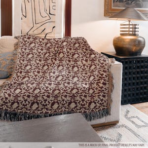May include: A rectangular throw blanket featuring a dark brown and cream woven pattern of stylised birds and intricate foliage. The blanket has a fringed edge along the bottom and is draped over a light-coloured sofa. Text reads: THIS IS A MOCK UP, FINAL PRODUCT RESULTS MAY VARY.