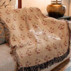 Op de afbeelding: Een beige sprei met een herhaald patroon van rode en bruine bloemmotieven. De deken heeft een gefranjerde rand in zwart en bruin. De sprei is over een bank gedrapeerd, voor de interieurdecoratie.