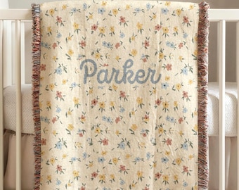 May include: A cream-colored baby blanket with a floral pattern in yellow, blue, and red. The name "Parker" is embroidered in blue script across the center. The blanket has a fringed edge.