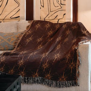 May include: A dark brown throw blanket with a repeating starburst pattern in a lighter brown color. The blanket is draped over a sofa and has a fringed edge. The throw is a decorative home textile.