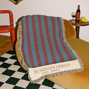 Pode incluir: Uma manta listrada em tons de azul e bordô com franjas, drapeada sobre uma poltrona amarela. A manta apresenta o texto "THE MADISON FAMILY EST. 2025" em uma borda creme. Um item decorativo para casa.