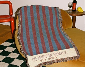 May include: A striped throw blanket in shades of blue and burgundy, draped over a yellow armchair. The blanket features the text "THE MADISON FAMILY EST. 2025" on a cream-colored border. The blanket has a fringed edge.