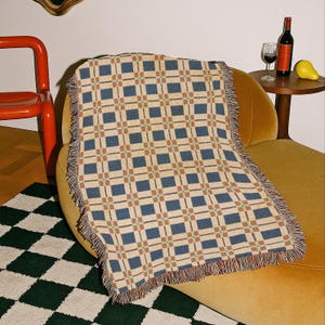 May include: A beige throw blanket with a blue and brown grid pattern and fringed edges. The blanket is draped over a yellow armchair. A red chair, a green and white checkered rug, and a small table with a bottle of wine and a pear are also visible.