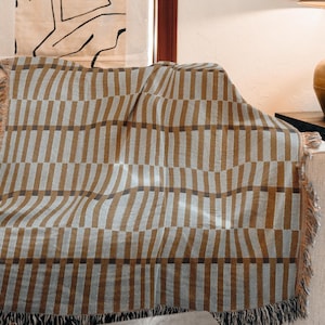 Op de afbeelding: Een bruin en grijs gestreepte plaid met franjes over een beige bank gedrapeerd. De deken heeft een herhalend verticaal streepjespatroon. De franjes zijn lichtbruin van kleur. Het is een decoratief huistextiel.