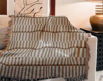 Puede incluir: Una manta a rayas marrones y grises con flecos, drapeada sobre un sofá beige. La manta presenta un patrón de rayas verticales repetidas. Los flecos son de color marrón claro. Es un textil decorativo para el hogar.
