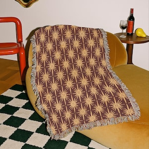 May include: A rectangular woven throw blanket featuring a repeating pattern of light tan starbursts on a dark brown background, finished with fringed edges along the sides. The text at the bottom reads: THIS IS A MOCK UP, FINAL PRODUCT RESULTS MAY VARY.