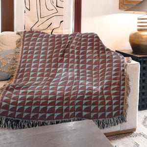 May include: A brown and blue geometric patterned throw blanket draped over a beige sofa. The blanket features a repeating pattern of half-circle shapes in a light blue color against a brown background. The blanket has a fringed edge.