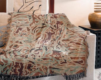 May include: A woven throw blanket with a camouflage pattern in shades of brown, green, and tan, draped over a beige sofa. The blanket has a fringed edge. Home decor item.