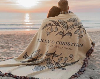 Pode incluir: Uma manta bege com um design floral e os nomes "KILEY & CHRISTIAN" e a data "NOVEMBER 22, 2025" impressos. A manta, com uma borda com franjas, está sobre duas pessoas numa praia ao pôr do sol.
