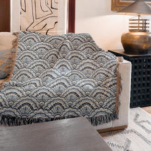 May include: A woven throw blanket featuring a repeating fan-shaped pattern in shades of navy blue, light blue, and cream. The blanket has a textured appearance with frayed edges along the sides and bottom, draped over a light-colored sofa.