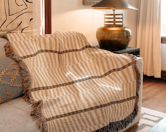 Puede incluir: Una manta a rayas beige y marrón con detalles de flecos sobre un sofá. La manta tiene un patrón repetitivo de rayas verticales en diferentes tonos de beige y marrón. Un cojín decorativo es visible en el sofá. Una lámpara de latón está sobre una mesa auxiliar.