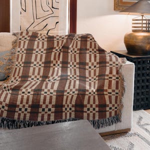 May include: A brown and beige woven throw blanket with a geometric pattern draped over a sofa. The blanket has a fringe along the edges and a mix of dark brown, beige, and black squares and rectangles. The throw blanket is a home decor item.