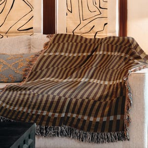 May include: A brown and tan striped throw blanket with fringe detail draped over a beige sofa. A decorative grey cushion with gold embroidery is visible. The blanket's pattern is a series of vertical stripes.