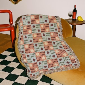 Könnte beinhalten: Eine gewebte Wohndecke mit einem sich wiederholenden Gittermuster aus abwechselnden Quadraten. Das Design umfasst hellblaue Quadrate mit dunkelbraunen Sternmotiven und pfirsichfarbene Quadrate mit grünen Rautenmustern, abgeschlossen mit Fransen.